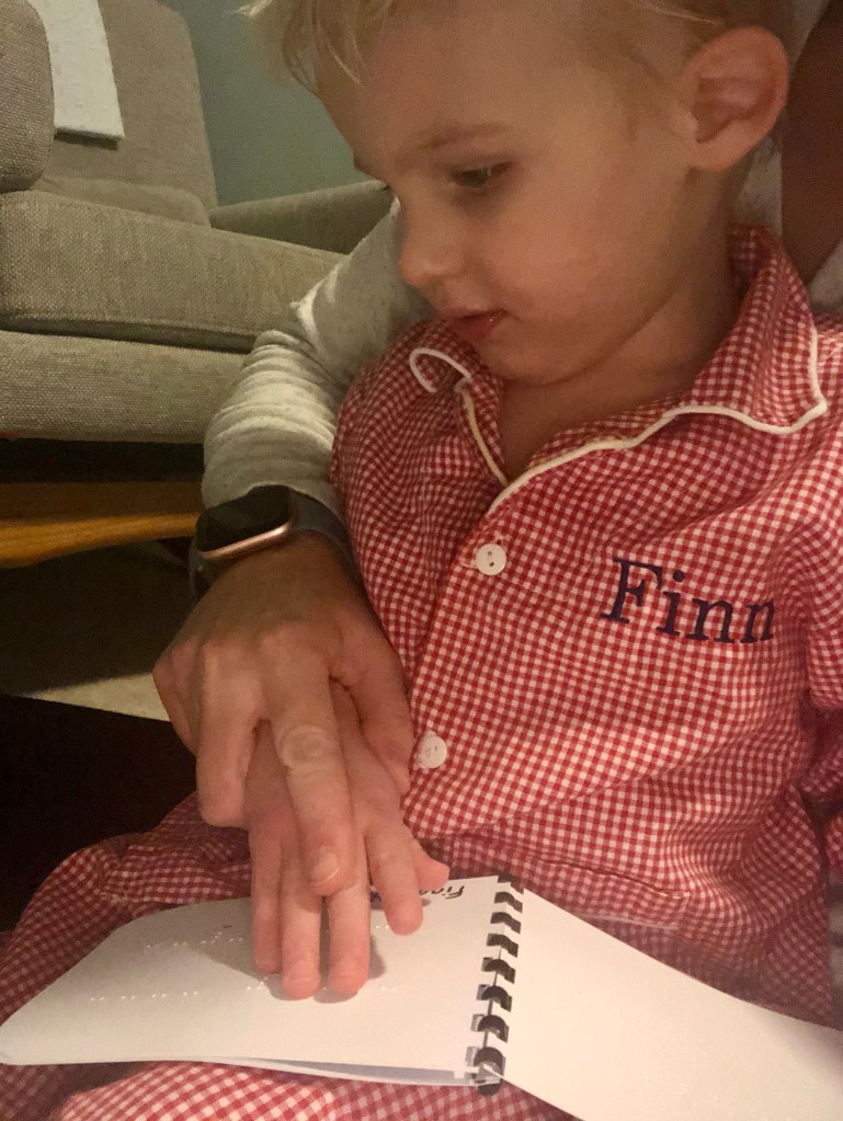 Little boy with his hand on a small brailled book. His mother has her hand over his guiding his fingers over the letters. 