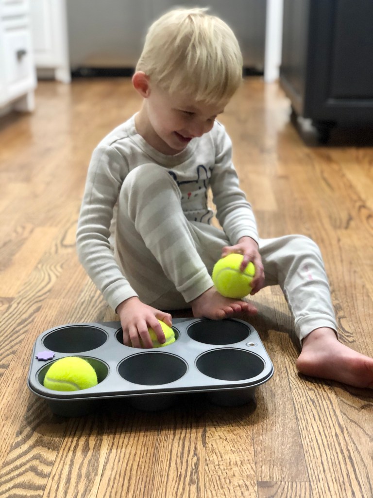 Little boy sitting on the kitchen floor with one hand holding a tennis ball. The other hand rests on a ball inside of a muffin tin. There is a third ball in the tin as well. 